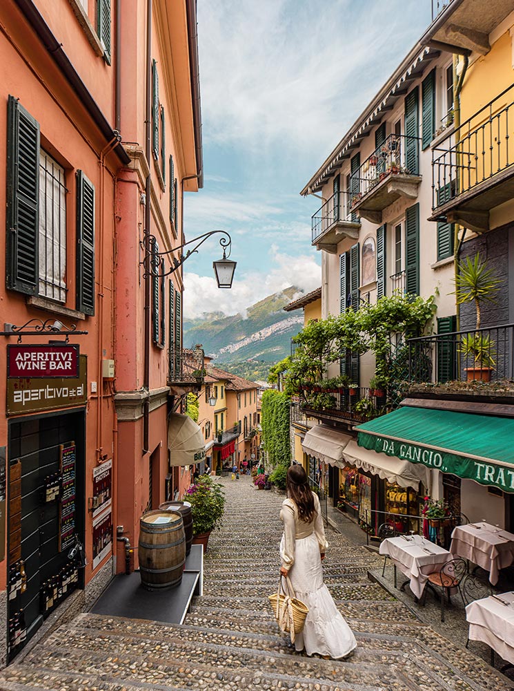 A woman in a flowing white dress walks down a charming cobblestone street in Bellagio, Lake Como, Italy. The picturesque alleyway is lined with colorful buildings, wine bars, and trattorias, with lush greenery and balconies overlooking the descending steps. In the background, the mountains and lake create a breathtaking view.