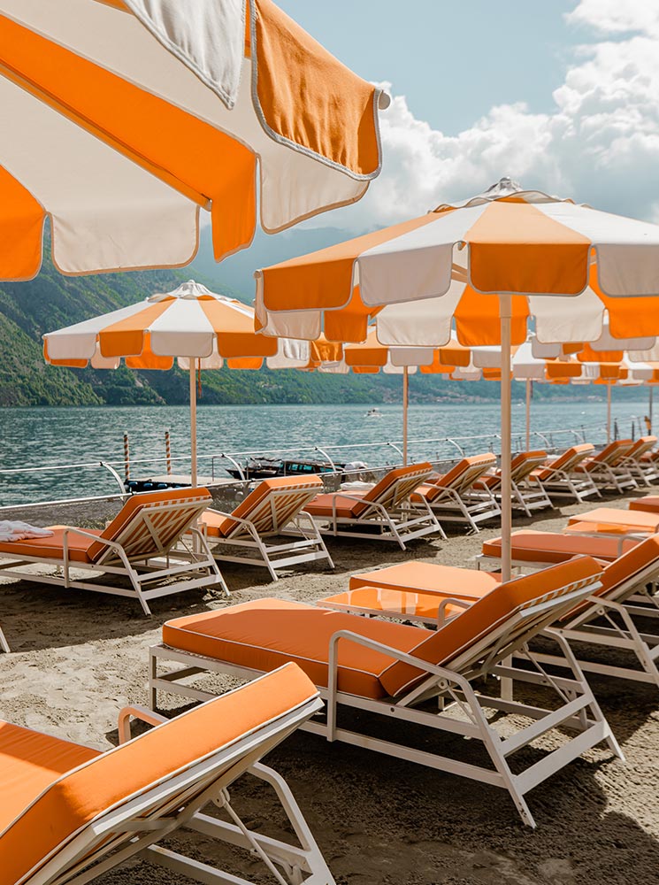 A luxurious beach club at Lake Como, Italy, featuring rows of orange and white striped umbrellas shading plush sun loungers on a sandy shoreline. The lake’s calm waters and lush green mountains provide a stunning backdrop, creating an inviting setting for relaxation. A towel draped over an umbrella adds to the laid-back, resort-style ambiance.