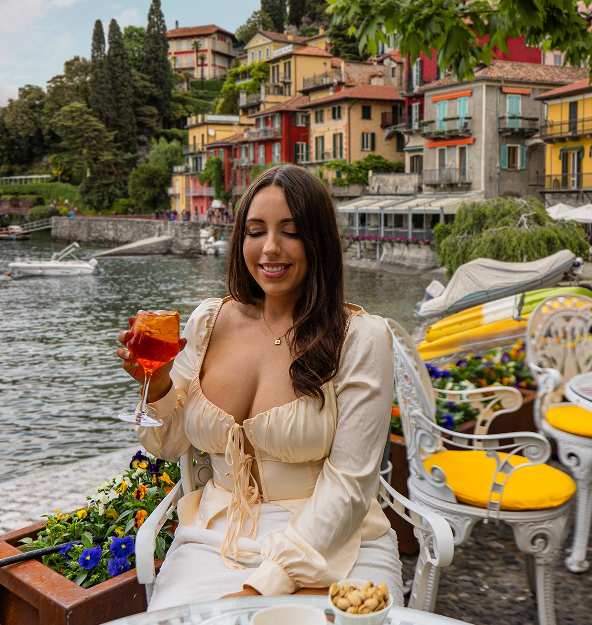 A woman in a cream-colored dress enjoys an Aperol Spritz at Bar Il Molo, a lakeside café in Varenna, Lake Como, Italy. She sits at a white ornate chair with yellow cushions, surrounded by colorful flowers, with the vibrant village buildings and calm waters of the lake in the background. Kayaks and boats are docked along the shore, adding to the charming waterfront ambiance.