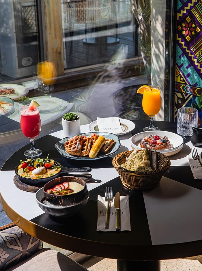 A beautifully arranged brunch spread at Boho Social in Doha, Qatar, features golden waffles, shakshuka, fresh fruit bowls, and a decadent dessert, accompanied by vibrant juices and a basket of warm flatbread. Sunlight streams through the window, highlighting the colorful and inviting presentation. The stylish table setting and cozy atmosphere enhance the relaxed yet upscale dining experience.