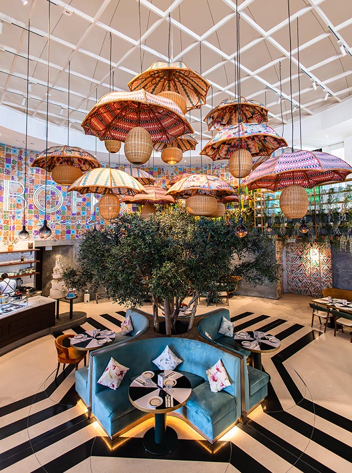 Boho Social in Doha, Qatar, features an eclectic and vibrant interior with colorful patterned umbrellas hanging from the ceiling, woven lanterns, and a central tree surrounded by plush blue seating. The bold black-and-white striped floor contrasts with the warm tones of the decor, creating a lively yet cozy dining atmosphere. The mix of bohemian and contemporary design elements enhances the restaurant's unique and artistic charm.