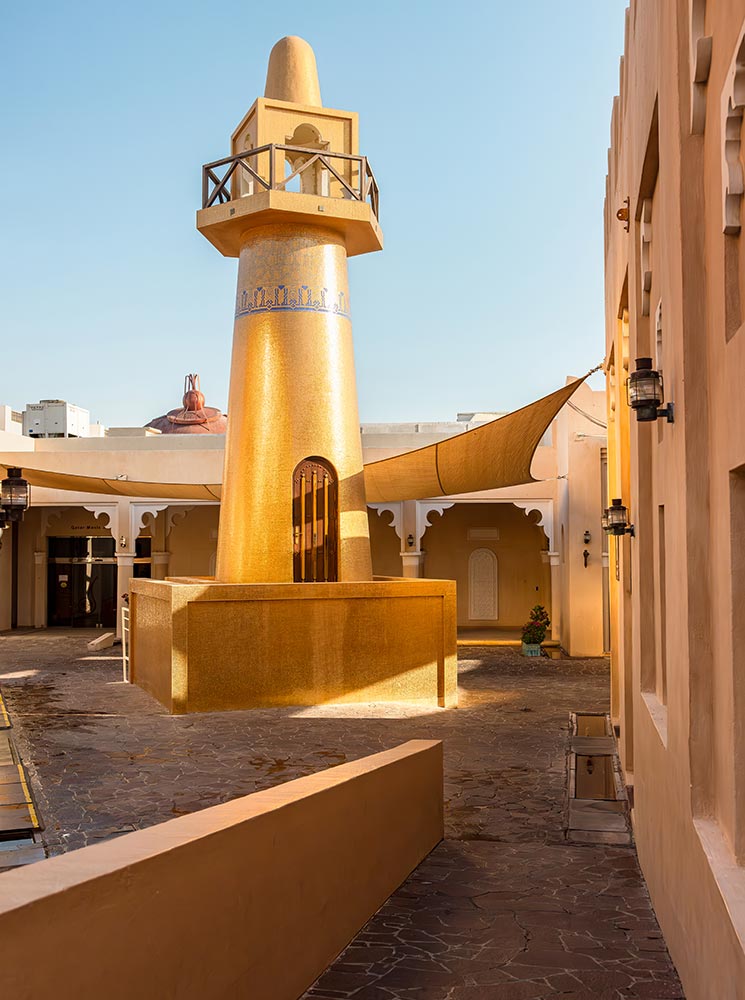 The golden mosque at Katara Cultural Village in Doha, Qatar, shines under the sunlight, featuring a striking minaret adorned with intricate blue tile detailing. The mosque's warm golden exterior contrasts with the surrounding traditional architecture and white archways. Soft shadows and warm tones create a serene and inviting atmosphere in this cultural and spiritual landmark.