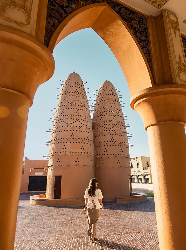 A woman walks barefoot towards the traditional pigeon towers at Katara Cultural Village in Doha, Qatar, framed by an ornate archway. The terracotta-colored towers, adorned with small holes and wooden perches, stand against a clear blue sky, reflecting traditional Qatari architecture. The warm lighting and surrounding historic buildings create a serene and culturally rich atmosphere.