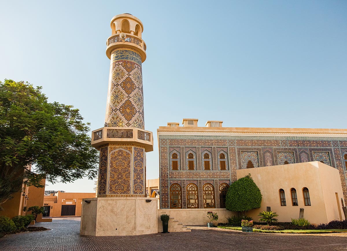 A beautifully designed minaret and mosque at Katara Cultural Village in Doha, Qatar, adorned with intricate Persian-style mosaic tiles in blue, gold, and brown hues. The surrounding architecture features traditional arched windows and geometric patterns, reflecting Islamic artistic heritage. The warm sunlight and lush greenery enhance the serene and historic atmosphere of this cultural landmark.







