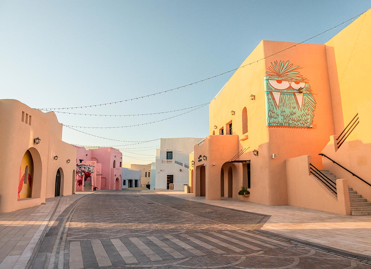 A colorful street in the Mina District of Doha, Qatar, features pastel-hued buildings with arched doorways, vibrant murals, and artistic graffiti. String lights hang above the cobblestone walkway, enhancing the charming and lively atmosphere. The warm glow of the setting sun casts soft shadows, adding to the district's inviting and creative ambiance.







