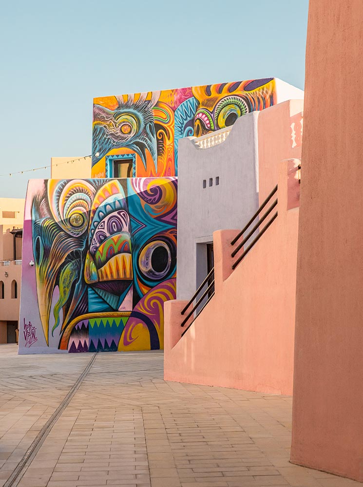 A vibrant mural featuring abstract and surreal designs in bold colors adorns the walls of pastel-colored buildings in the Mina District of Doha, Qatar. The intricate artwork contrasts with the soft hues of the surrounding architecture, creating a striking visual display. The empty walkway and warm sunlight enhance the artistic and cultural charm of this creative neighborhood.