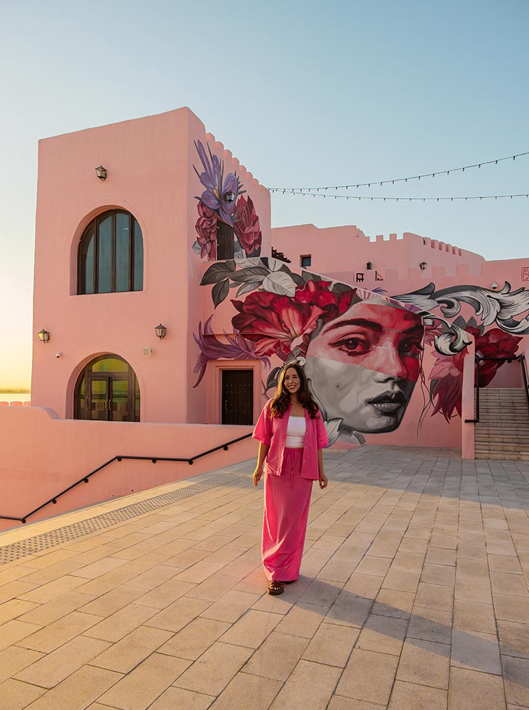 A woman dressed in a vibrant pink outfit walks through the Mina District in Doha, Qatar, with a striking mural of a woman's face adorned with floral artwork on a pastel pink building behind her. The warm glow of the setting sun casts a golden light, enhancing the artistic and modern aesthetic of the area. String lights above and the architectural details add to the district’s creative and inviting atmosphere.