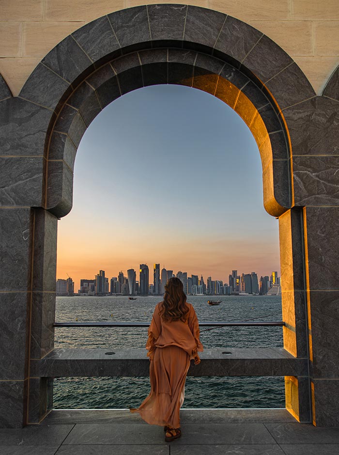 A woman in a flowing brown dress stands framed by a grand stone archway at the Museum of Islamic Art in Doha, Qatar, gazing at the city skyline across the water. The sunset casts a golden glow on the scene, illuminating the modern high-rises and traditional wooden dhows in the bay. The moment captures the contrast between Qatar’s rich history and its futuristic urban landscape.