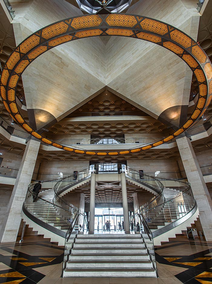 The grand interior of the Museum of Islamic Art in Doha, Qatar, features a sweeping staircase with glass railings leading to multiple levels. A striking geometric chandelier encircles the ceiling, casting warm light onto the surrounding limestone walls. The intricate architectural details reflect Islamic design elements, creating a space that is both modern and steeped in cultural heritage.