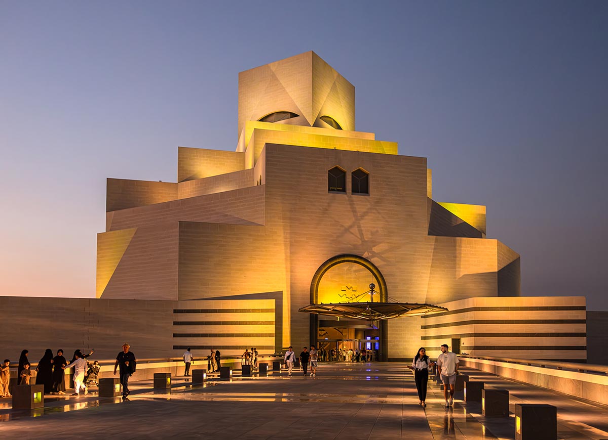 The Museum of Islamic Art in Doha, Qatar, is illuminated against the evening sky, showcasing its striking geometric design inspired by traditional Islamic architecture. Visitors walk along the sleek, reflective promenade leading to the entrance, where golden lighting accentuates the building's angular structure. The modern yet timeless aesthetic of the museum makes it a cultural landmark on the Doha waterfront.






