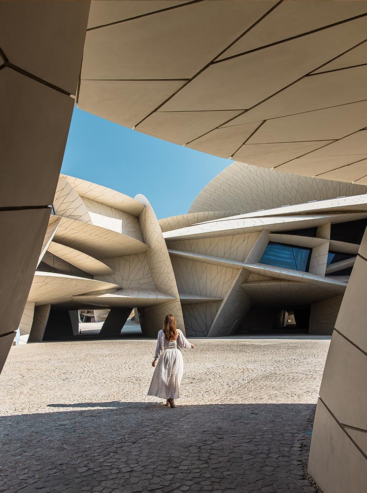 A woman in a flowing white dress walks toward the National Museum of Qatar in Doha, surrounded by its striking desert rose-inspired architecture. The building's interlocking geometric panels create a futuristic yet organic design, with soft sunlight casting shadows on the textured ground. The scene captures the blend of innovation and cultural heritage that defines this iconic museum.