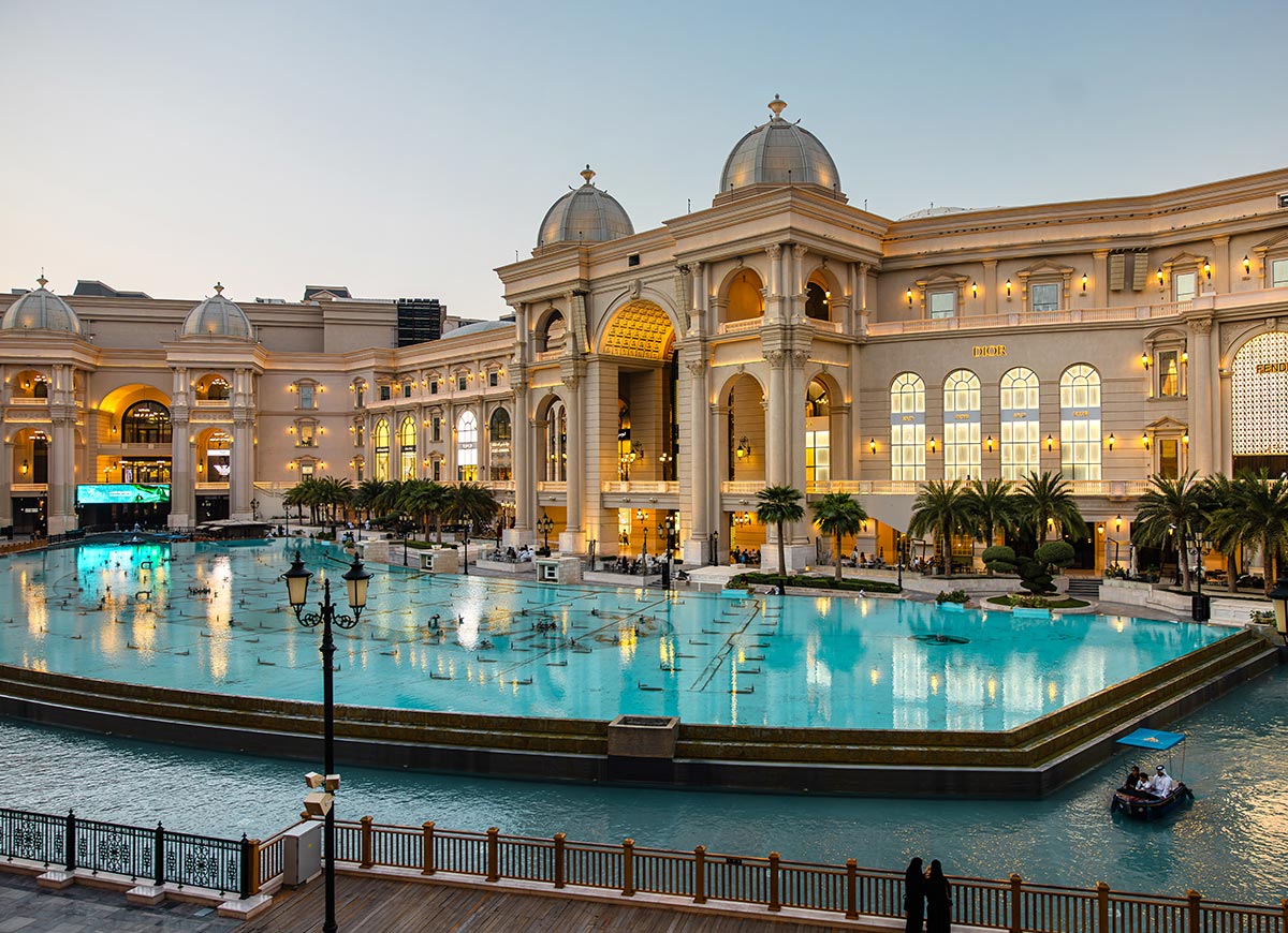 The grand exterior of Place Vendôme in Doha, Qatar, showcases its opulent European-inspired architecture with domed rooftops, towering archways, and elegant storefronts. A large turquoise water feature with fountains reflects the golden lights of the luxury shopping complex, while a small boat glides across the canal. The scene exudes sophistication, blending classical French design with modern Qatari elegance.






