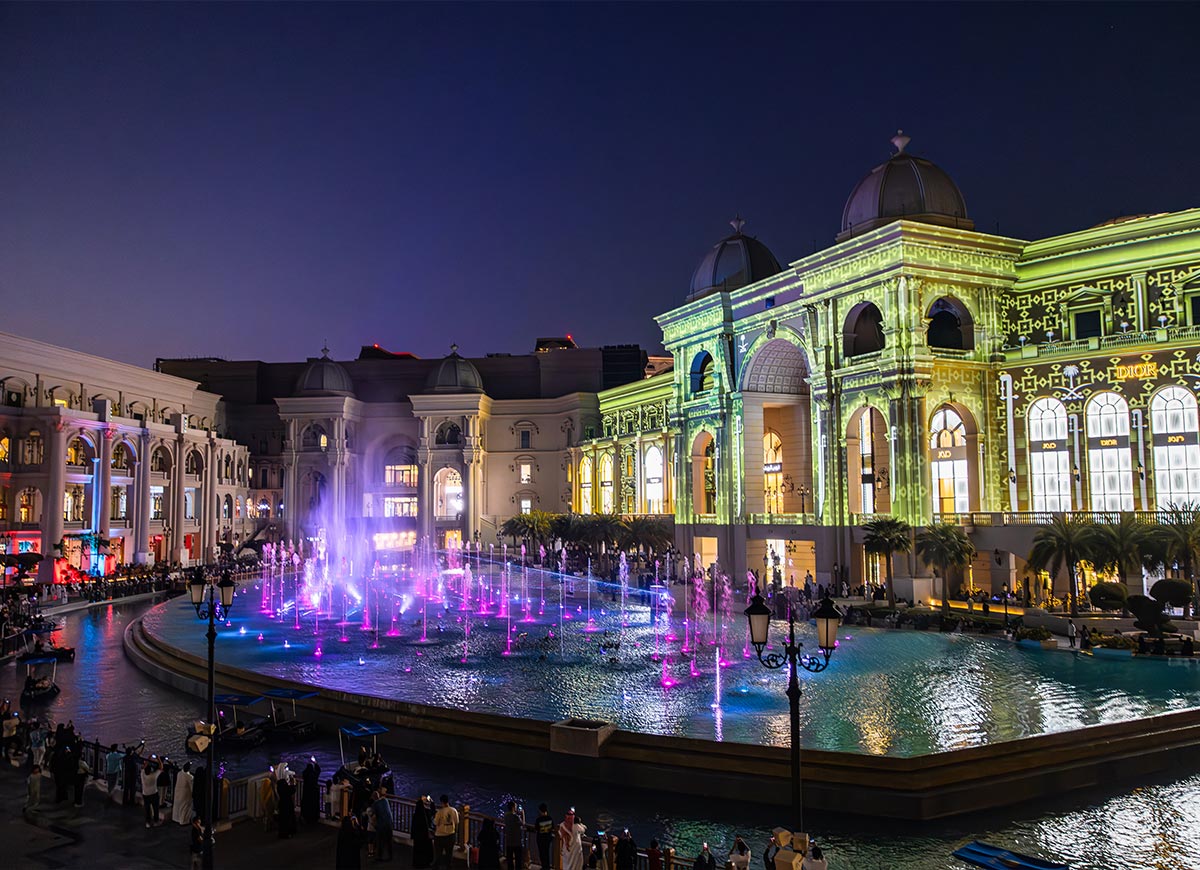 The Place Vendôme fountain show in Doha, Qatar, dazzles at night with vibrant pink and purple water jets synchronized to music. The luxurious shopping complex's grand façade is illuminated with intricate green and gold projections, enhancing its European-inspired architecture. Crowds gather along the waterfront promenade to enjoy the mesmerizing display of lights, water, and music in this elegant setting.







