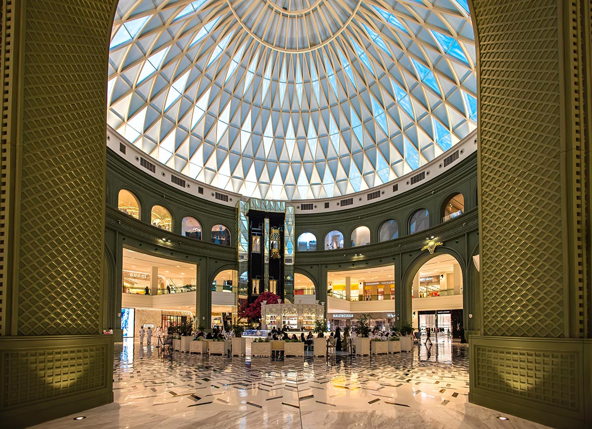 The grand interior of Place Vendôme shopping center in Doha, Qatar, features a stunning glass-domed ceiling allowing natural light to illuminate the luxurious space. Elegant green archways, marble flooring, and high-end designer boutiques like Balenciaga and Gucci create an opulent shopping experience. A stylish central café with lush floral decor adds to the sophisticated ambiance of this architectural masterpiece.







