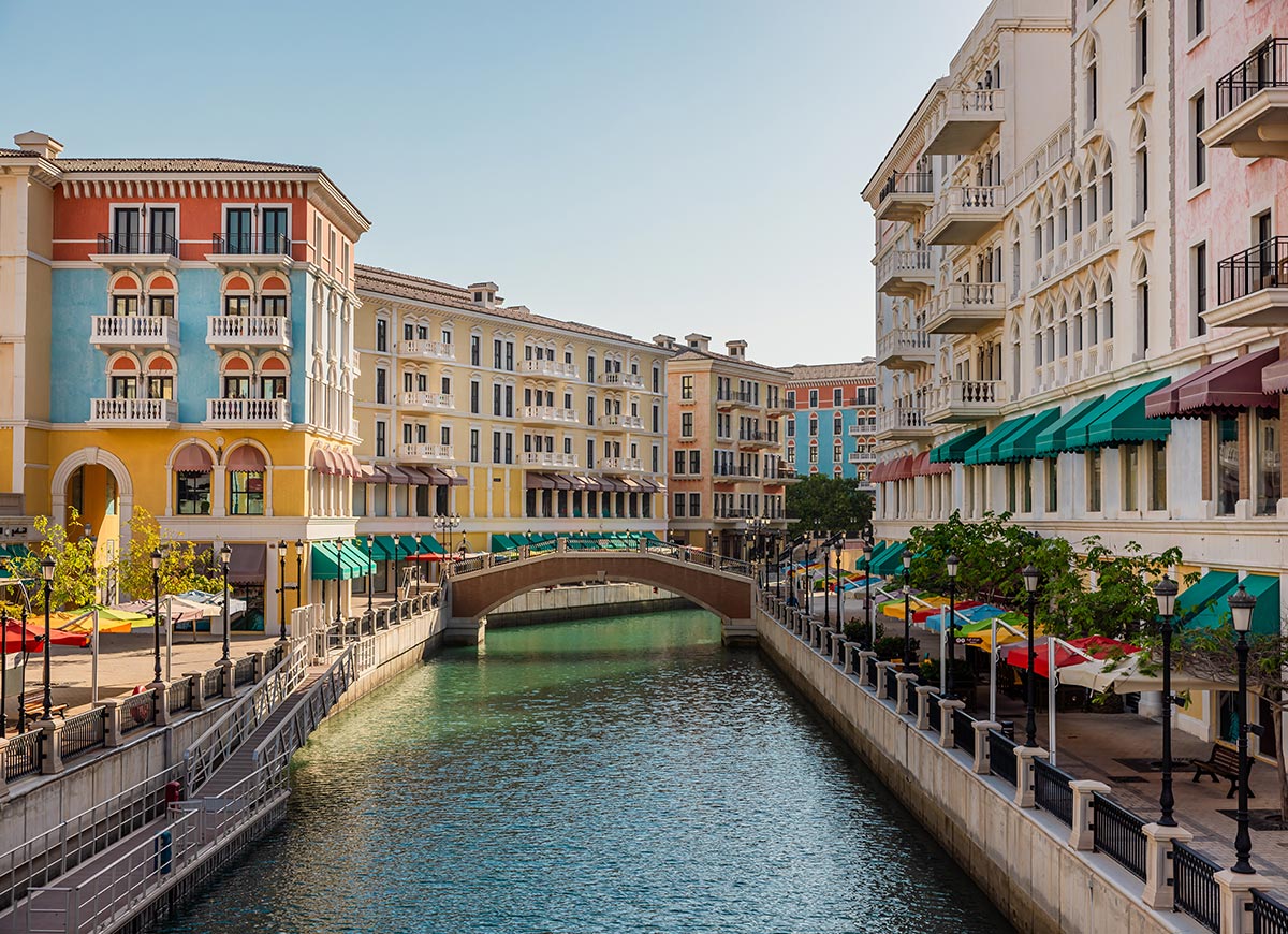 A picturesque canal in Qanat Quartier, Doha, Qatar, is lined with pastel-colored Venetian-style buildings featuring balconies, arched windows, and striped awnings. A charming stone bridge spans the turquoise water, while waterfront cafés with colorful umbrellas add a lively European ambiance. The peaceful scene reflects the unique blend of Qatari luxury and Mediterranean charm.







