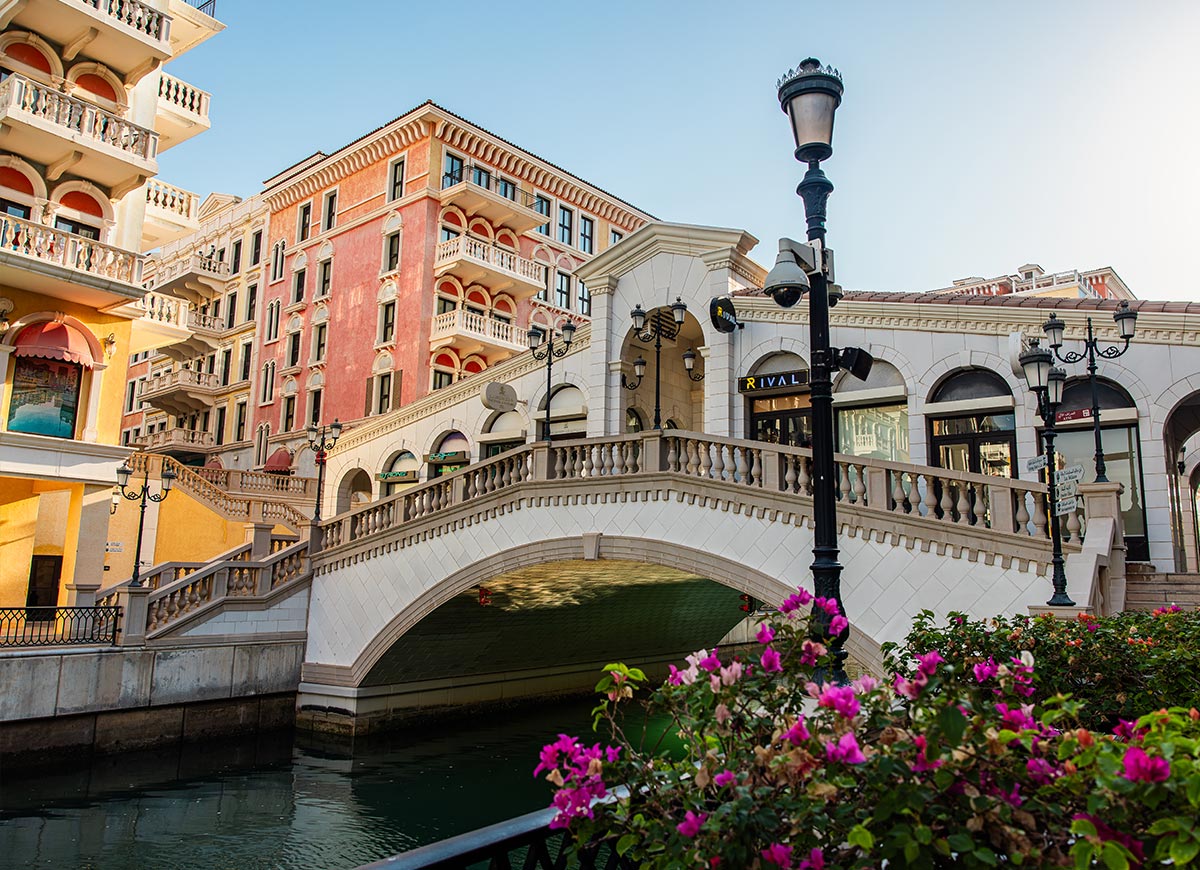 A charming Venetian-style bridge arches over a canal in Qanat Quartier, Doha, Qatar, surrounded by pastel-colored buildings with elegant balconies and arched windows. The warm sunlight enhances the Mediterranean-inspired architecture, while vibrant pink flowers in the foreground add a touch of natural beauty. The peaceful waterfront setting exudes a European ambiance in the heart of the Middle East.







