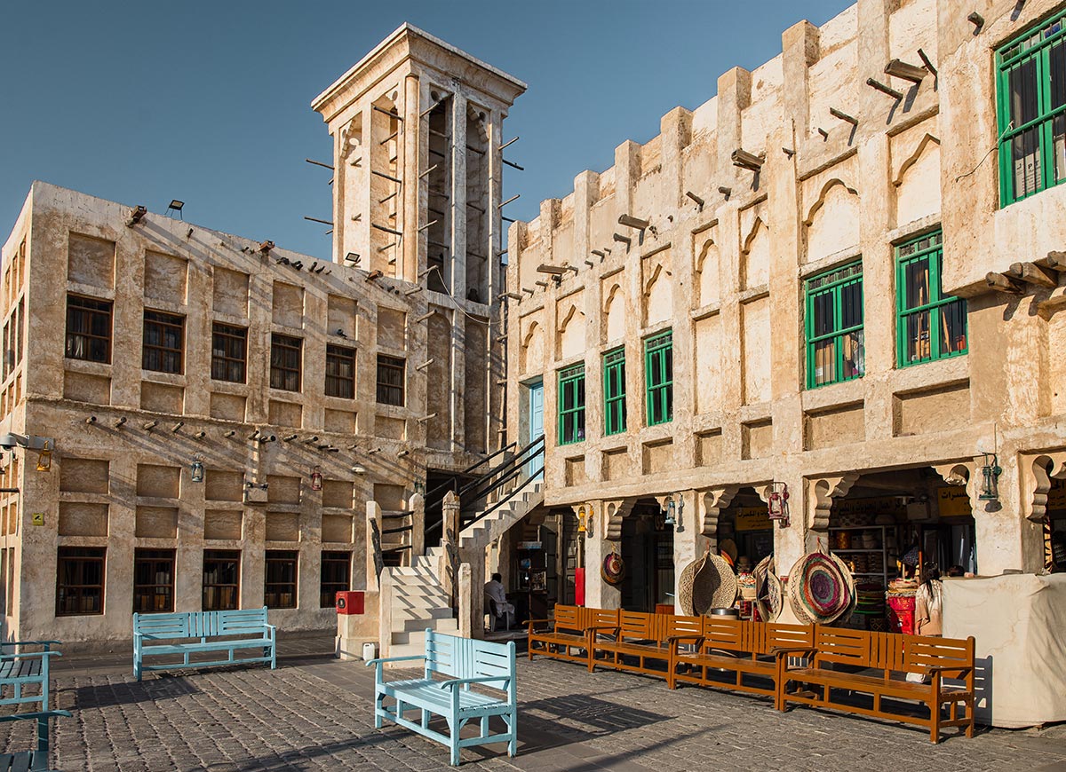 A traditional marketplace in Souq Waqif, Doha, Qatar, features historic beige-colored buildings with wooden beams, arched windows, and vibrant green shutters. The open courtyard is lined with wooden benches and pastel blue seating, while shops display handmade crafts, woven baskets, and local goods. The sun casts warm light over the scene, highlighting the souq’s cultural charm and authentic Arabian ambiance.






