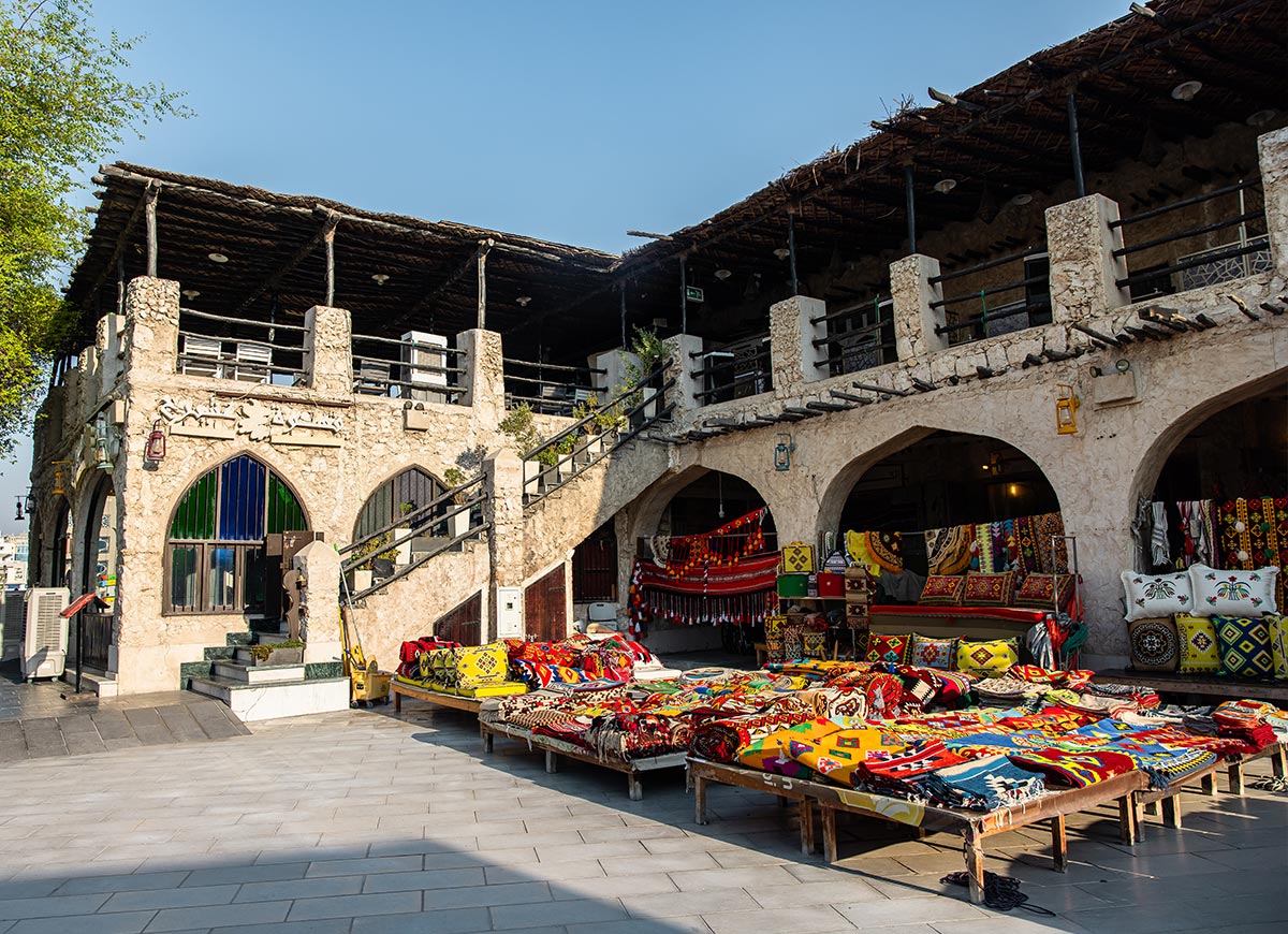 A traditional marketplace in Souq Waqif, Doha, Qatar, showcases a rustic stone building with arched doorways, wooden railings, and a shaded upper-level terrace. Vibrant, handcrafted textiles, cushions, and rugs in bold patterns and rich colors are displayed on wooden benches in the open courtyard. The warm sunlight enhances the lively and authentic atmosphere of this historic market.







