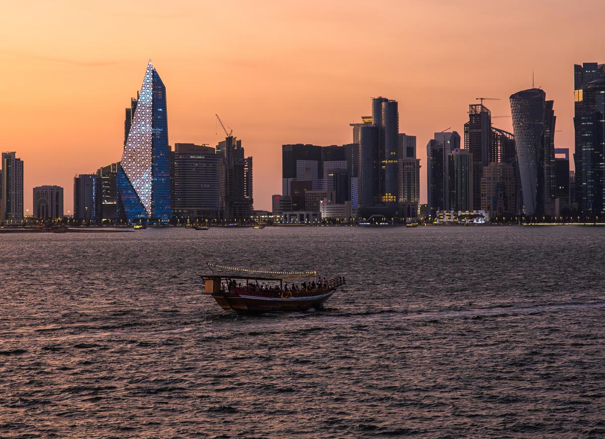 A traditional dhow boat sails across the waters of Doha, Qatar, at sunset, with the illuminated city skyline in the background. The sky glows in warm orange hues, reflecting off the modern skyscrapers, including the uniquely designed glass tower. The peaceful scene captures the blend of Qatar’s maritime heritage and contemporary urban elegance.