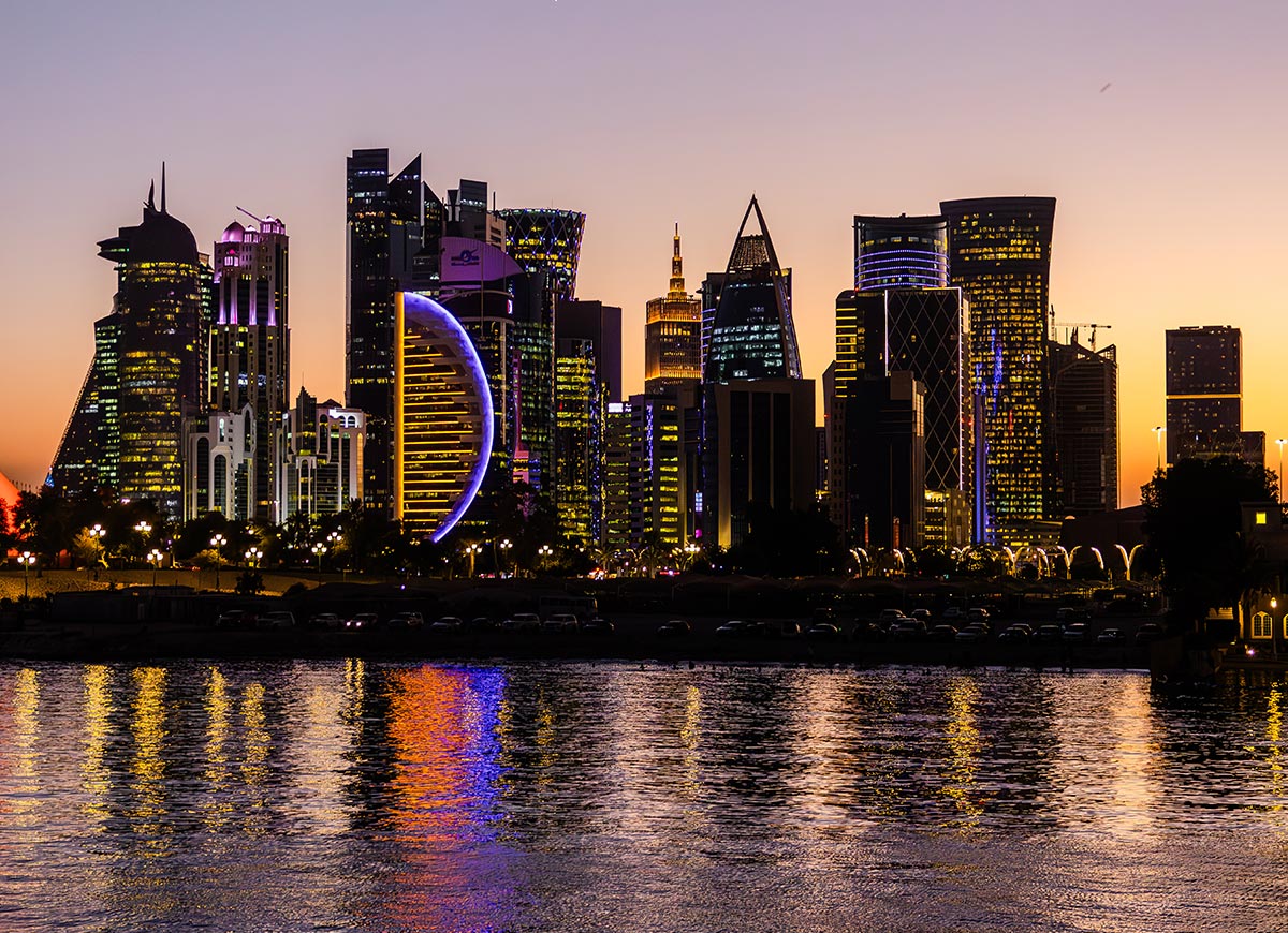 A breathtaking view of the Doha city skyline at sunset, with the sky transitioning from warm golden hues to deep twilight tones. The illuminated skyscrapers reflect off the calm waterfront, creating a colorful and dynamic cityscape. The iconic, crescent-shaped tower stands out with its vibrant lighting, adding to the futuristic charm of Qatar's capital.







