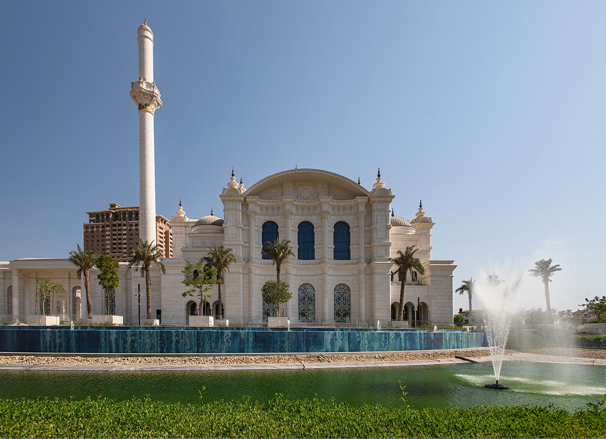 A grand white mosque in Doha, Qatar, stands against a clear blue sky, featuring elegant domes, arched windows, and a towering minaret. A lush green landscape with a reflective water feature and fountains enhances the serene and majestic ambiance. The intricate architectural details and palm-lined surroundings create a striking blend of tradition and luxury.






