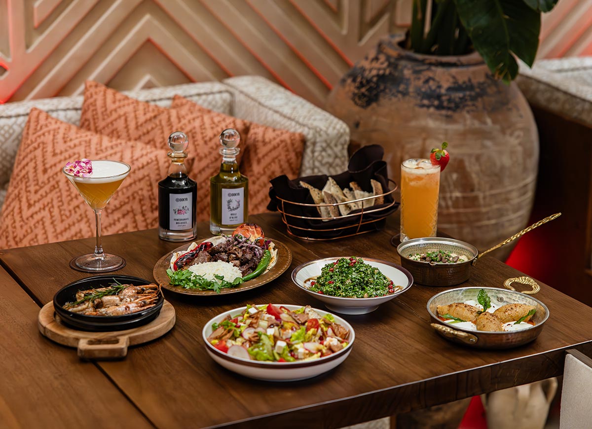 A beautifully arranged Middle Eastern feast is set on a wooden table at a restaurant in Doha, Qatar, featuring dishes like tabbouleh, grilled shrimp, hummus, fresh salad, and kibbeh. Two elegant cocktails, a basket of artisanal bread, and bottles of flavored oil and vinegar accompany the meal. The warm ambiance, with patterned cushions and rustic decor, enhances the luxurious dining experience.







