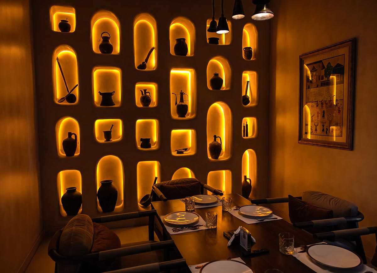 A cozy dining space in a Doha restaurant features a warmly lit wall with arched niches displaying traditional pottery and antique artifacts. The soft amber lighting creates an intimate and inviting ambiance, complementing the dark wood dining table set with elegant tableware. A framed artwork on the wall adds a cultural touch to the rustic yet refined setting.







