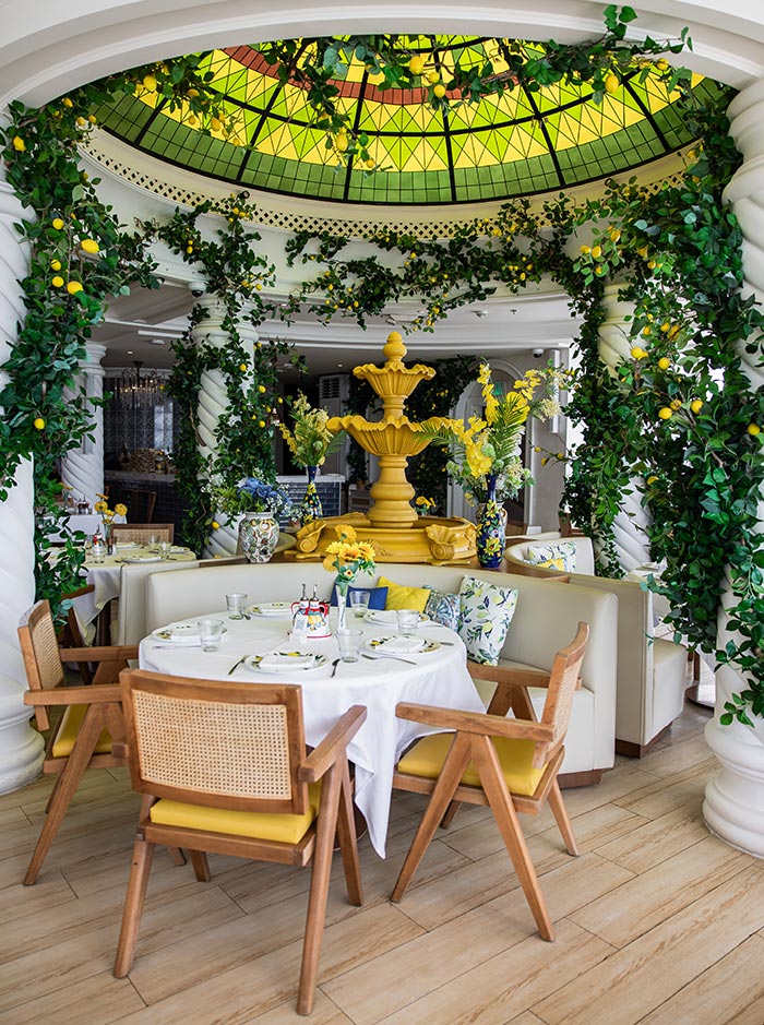 A beautifully designed dining space at Lucia’s Italian restaurant in Doha, featuring a Mediterranean-inspired setting with lush greenery and lemon trees. A stained-glass dome casts a warm glow over the yellow centerpiece fountain, surrounded by elegant wooden chairs and cozy white booths. The tables are set with fine dining details, evoking the charm of the Amalfi Coast in a luxurious yet inviting ambiance.