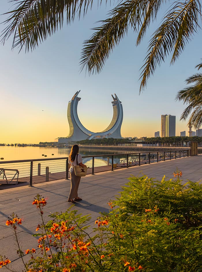 A woman walks along a waterfront promenade in Doha, Qatar, gazing at the striking Katara Towers in the distance. The scene is bathed in warm golden light, with palm fronds and vibrant orange flowers framing the foreground. The calm water reflects the towers’ unique curved design, creating a serene and picturesque moment.