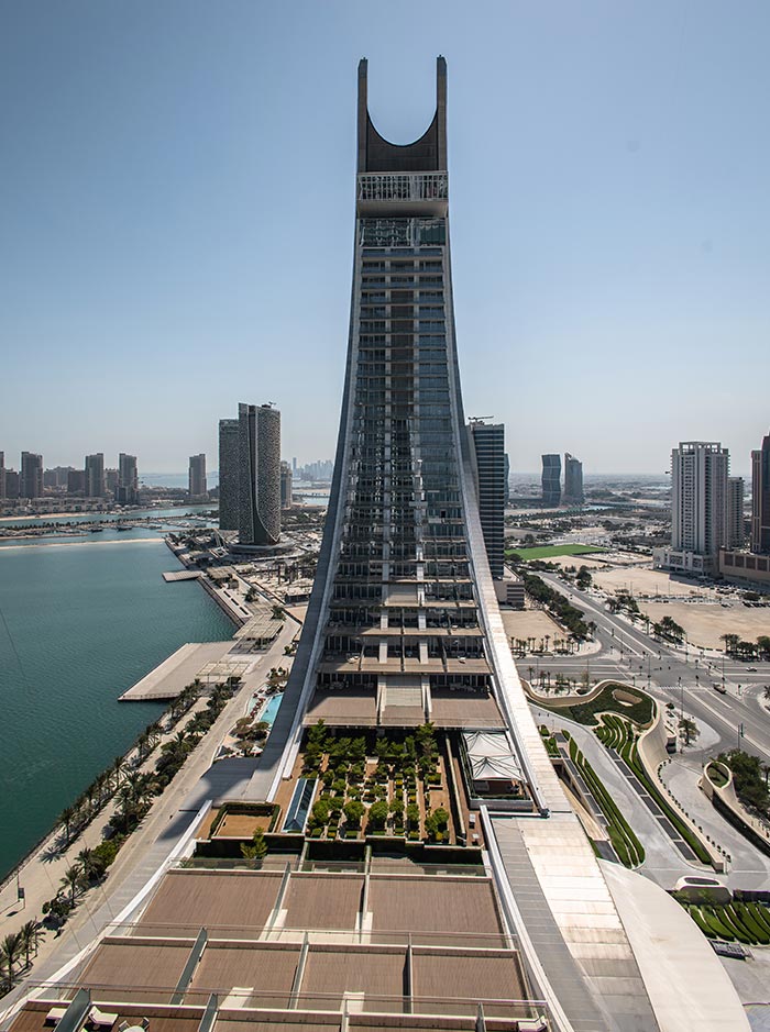 A striking aerial view of Katara Towers in Doha, Qatar, showcasing its unique, curved architectural design. The symmetrical structure rises above the cityscape, overlooking the waterfront and surrounding modern high-rises. The foreground features a beautifully landscaped terrace, while the blue waters of the Arabian Gulf stretch beyond the city.