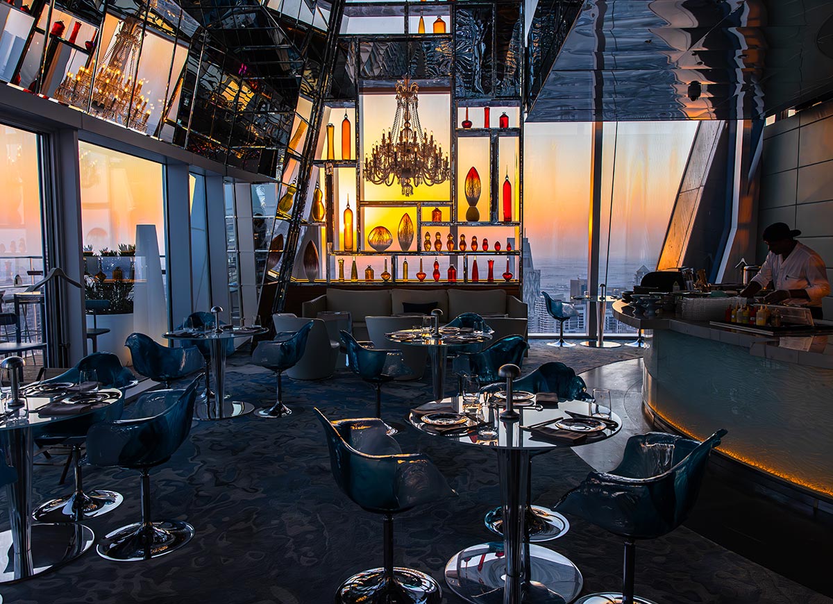 A luxurious rooftop bar in Doha, Qatar, set inside Katara Towers, offers a stylish ambiance with a backdrop of a golden sunset. The interior features sleek glass tables, modern blue chairs, and a striking illuminated bar adorned with decorative bottles and a grand chandelier. Large windows and mirrored surfaces enhance the panoramic cityscape views, creating an upscale and sophisticated atmosphere.







