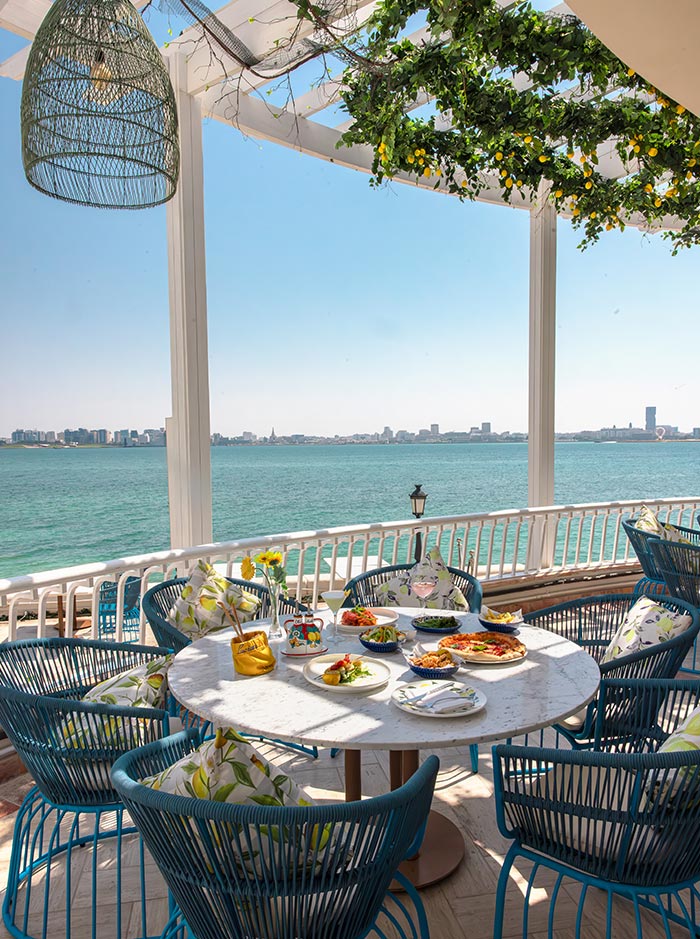 A picturesque dining setup at Lucia’s Italian restaurant in Doha, overlooking the sparkling blue waters with a city skyline in the distance. The round marble table is adorned with a vibrant Mediterranean feast, featuring fresh salads, pasta, and appetizers. The stylish blue chairs, floral cushions, and greenery-draped pergola create a charming, coastal-inspired ambiance perfect for an alfresco meal.