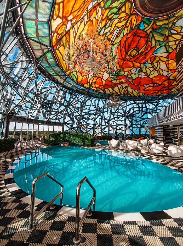 The rooftop pool at Mondrian Doha features a striking stained-glass dome ceiling with vibrant floral patterns, casting colorful reflections on the water. A grand crystal chandelier hangs in the center, adding a touch of opulence to the modern, geometric glass structure. The black-and-white checkered flooring and sleek white lounge chairs create a luxurious and artistic ambiance.