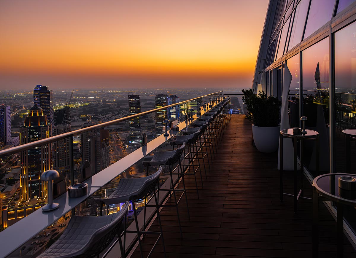 A sleek rooftop bar in Doha, Qatar, offers a breathtaking panoramic view of the city skyline at sunset. The warm orange hues of the sky fade into the evening lights of the towering skyscrapers below. Rows of stylish barstools line the glass railing, inviting guests to enjoy the elevated experience with a drink in hand.







