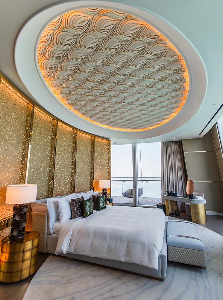 A luxurious hotel suite at Raffles Doha features a plush king-sized bed with elegant pillows, gold-accented side tables, and soft ambient lighting. The intricately designed ceiling has a wave-like pattern with warm backlighting, complementing the shimmering gold panel wall. A private balcony with hanging chairs offers a serene ocean view, enhancing the sophisticated and tranquil atmosphere.