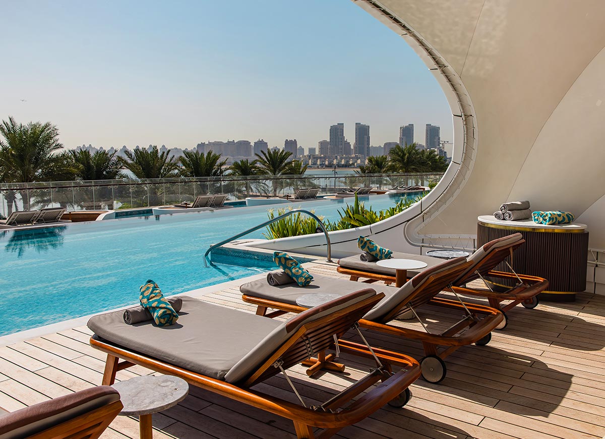 A luxurious rooftop infinity pool with wooden lounge chairs, neatly arranged with rolled towels and patterned cushions. The pool overlooks a city skyline with tall buildings in the background, surrounded by palm trees and a clear blue sky. A modern curved canopy adds a touch of elegance to the poolside setting, creating a tranquil and stylish retreat.







