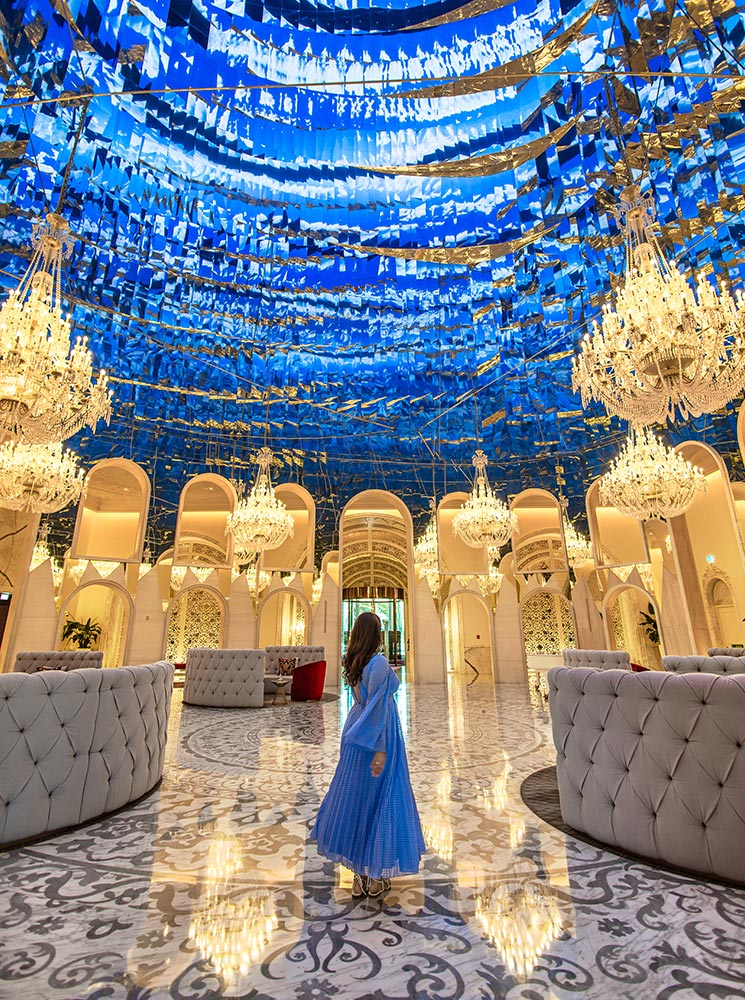 A woman in a flowing blue dress walks through the grand lobby of Raffles Doha, surrounded by opulent chandeliers, tufted seating, and intricate architectural details. The mirrored ceiling reflects vibrant blue hues, creating a mesmerizing, almost surreal atmosphere. The polished marble floor, elegant arches, and warm lighting enhance the luxurious ambiance of this breathtaking hotel lobby.