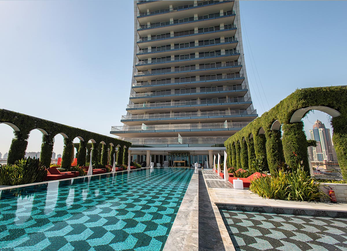 The rooftop pool area of Raffles Doha showcases a shimmering blue geometric-tiled pool, flanked by lush green archways and vibrant red lounge chairs. The hotel's modern, curved facade rises in the background, with the city skyline visible in the distance. The scene exudes luxury and tranquility, offering a serene oasis in the heart of Doha.







