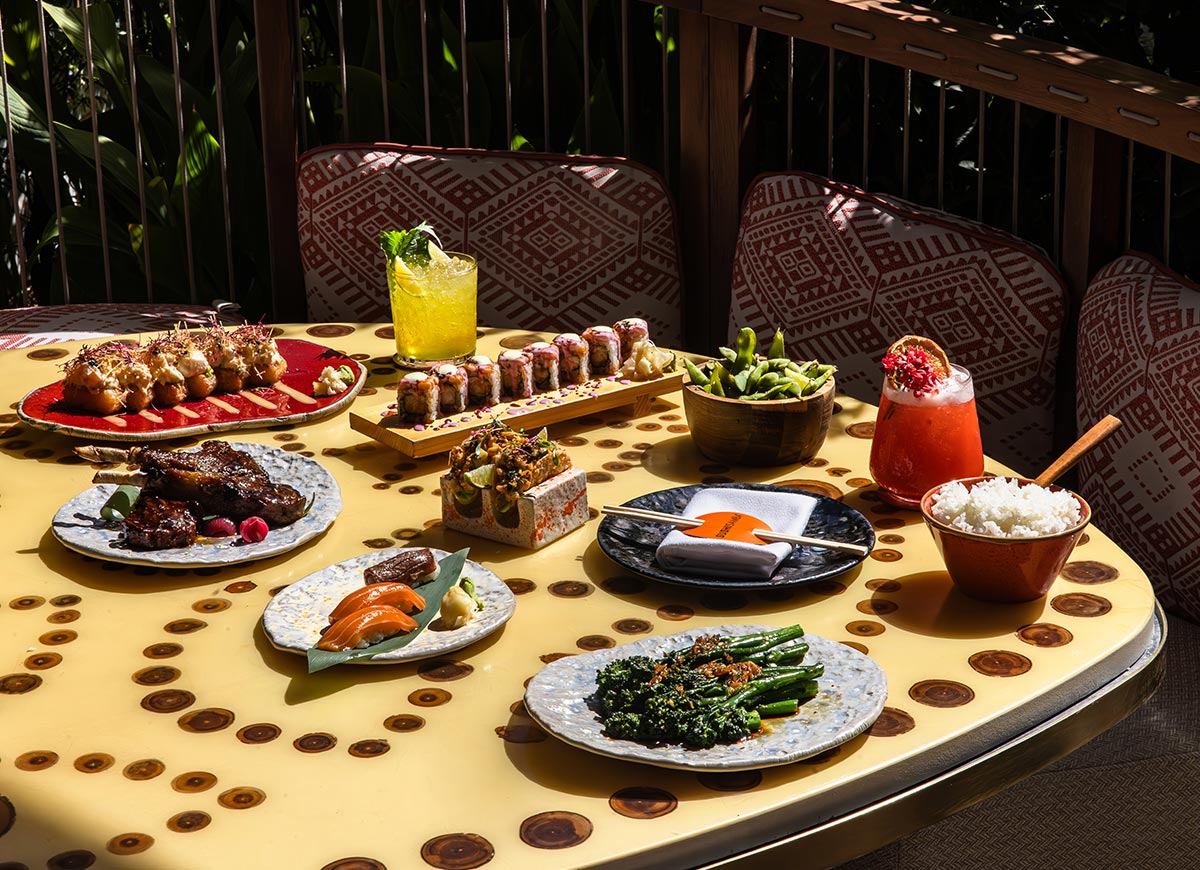 A beautifully arranged spread of Japanese and Peruvian fusion dishes is set on a vibrant outdoor table at SUSHISAMBA in Doha, Qatar. The feast includes sushi rolls, grilled lamb, salmon sashimi, broccolini, edamame, and a variety of colorful cocktails. The stylish seating with patterned cushions and warm natural lighting enhances the luxurious dining experience.







