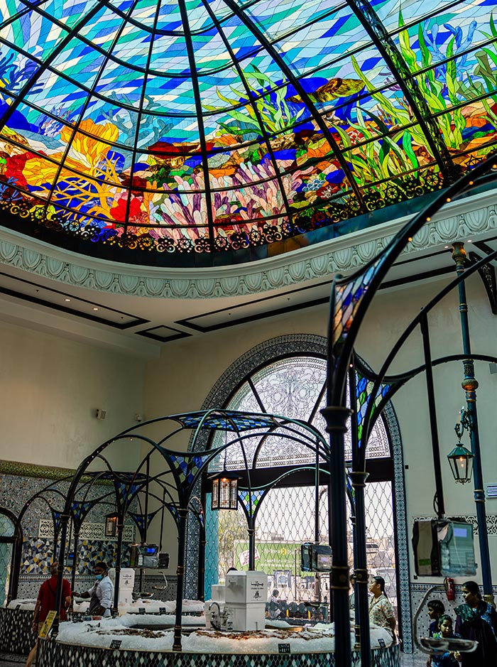 The Doha Fish Market in the Mina District showcases a stunning stained-glass dome ceiling with intricate underwater-themed artwork featuring coral reefs, fish, and sea plants. The elegant wrought-iron arches and mosaic-tiled counters create a sophisticated yet vibrant marketplace ambiance. Fresh seafood is displayed on beds of ice, while shoppers browse beneath the colorful, sunlit canopy.