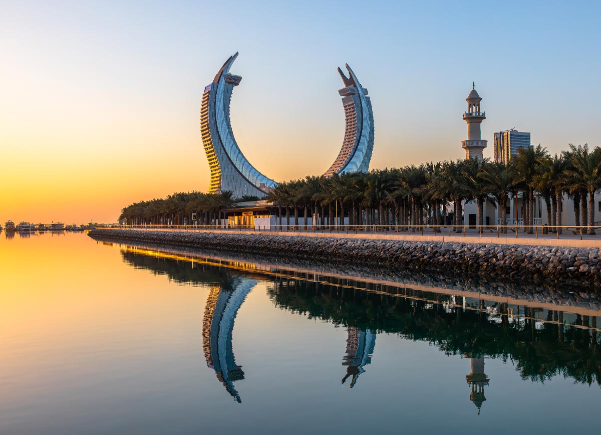 A stunning view of Katara Towers in Doha, Qatar, captured at sunrise, reflecting perfectly in the calm waters. The twin-arched skyscrapers, designed to resemble Qatar’s national emblem, stand against a golden sky, with palm-lined walkways and a mosque adding to the serene atmosphere. The image beautifully showcases the city's blend of modern architecture and cultural heritage.







