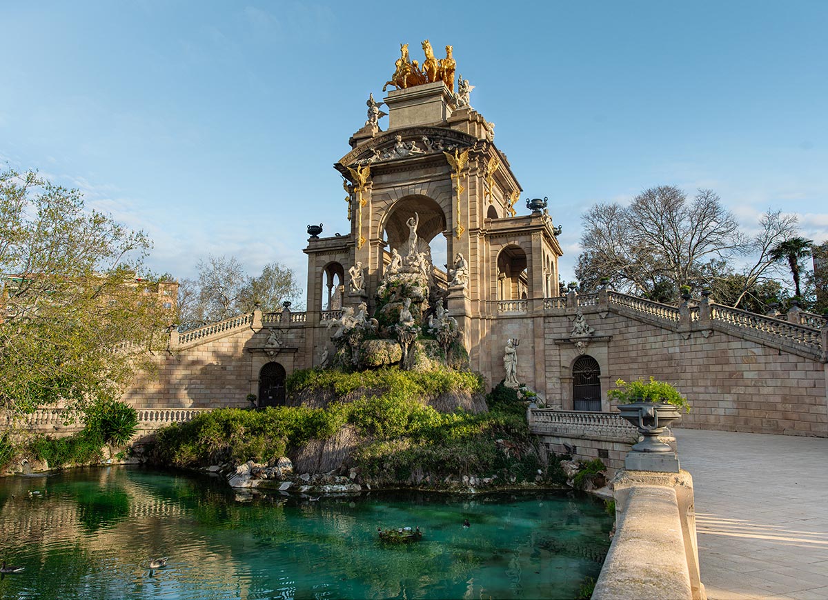 Ornate fountain at Ciutadella Park in Barcelona, featuring a grand stone monument with classical statues, cascading greenery, and golden sculptures of a chariot pulled by four horses at the top. The fountain sits above a serene turquoise pond surrounded by stone walkways and lush trees under a clear blue sky.