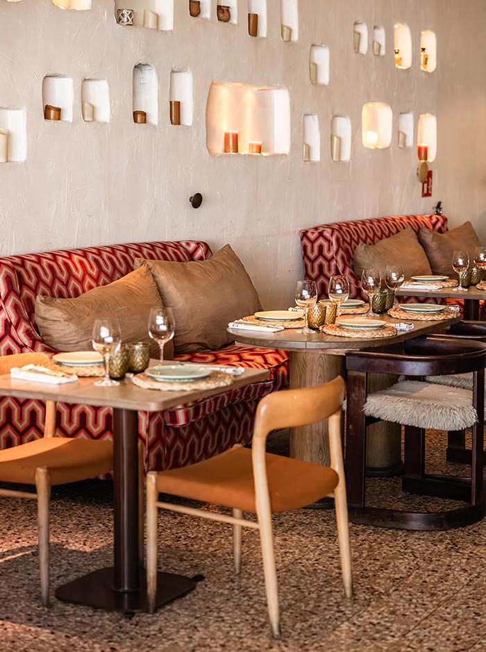 Chic interior of Julieta’s restaurant in Barcelona, with patterned red and beige banquettes, cozy earth-toned pillows, and modern wooden tables set with gold-toned glasses and light ceramic plates. The textured wall is adorned with softly lit alcoves holding candles, creating a warm, inviting atmosphere.