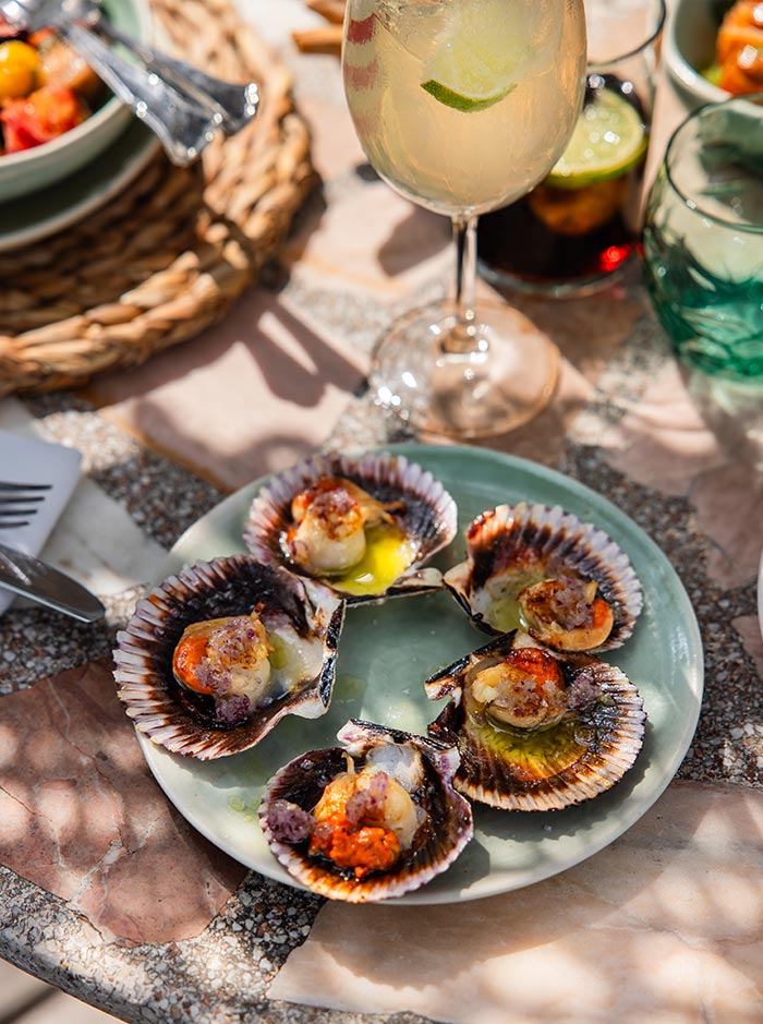 Sunlit table at Julieta’s restaurant in Barcelona, featuring a green ceramic plate of grilled scallops served in shell with melted butter and seasoning. A refreshing cocktail garnished with lime and additional drinks in the background complete the inviting Mediterranean setting.