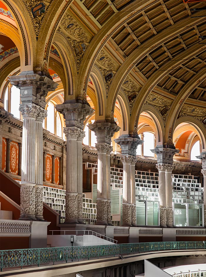 Interior detail of the Museu Nacional d'Art de Catalunya (MNAC) in Barcelona, featuring grand Roman-style columns with ornate carvings, a series of arched windows, and an elaborately decorated vaulted ceiling. The soft natural light highlights the intricate patterns and textures throughout the space.