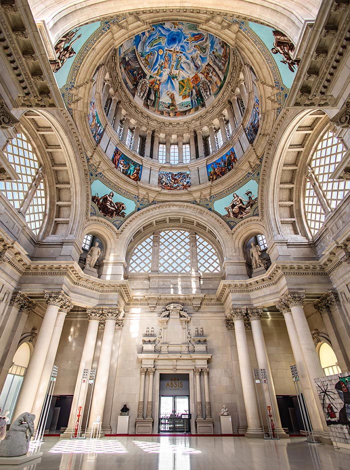 Interior view of the Museu Nacional d'Art de Catalunya (MNAC) in Barcelona, looking up at the grand dome adorned with colorful frescoes depicting historical and mythological scenes. The hall features neoclassical columns, arched windows with lattice patterns, and a central doorway labeled “ABSIS.”