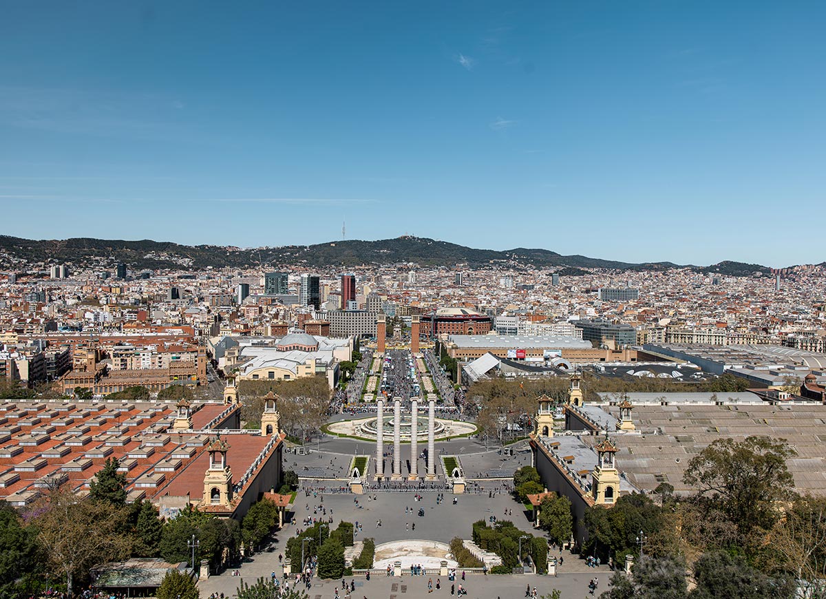 Panoramic view of Barcelona from the rooftop of the Museu Nacional d'Art de Catalunya (MNAC), looking down over Plaça d'Espanya with its twin Venetian towers, four white columns, and bustling avenues. The city stretches into the distance with a mix of historic and modern buildings backed by rolling hills.







