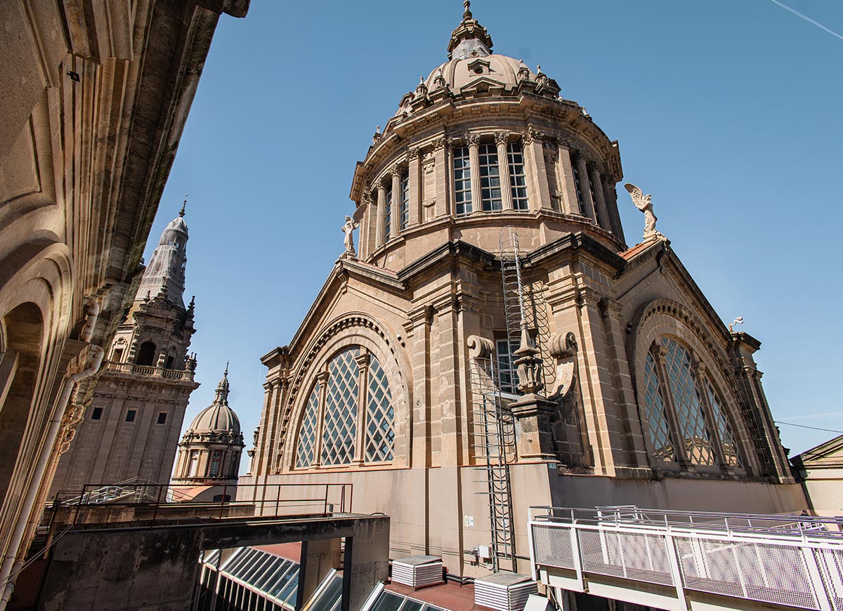 Rooftop view of the Museu Nacional d'Art de Catalunya (MNAC) in Barcelona, highlighting the grand dome with arched windows, classical columns, and winged statues. The architectural details are captured under a clear blue sky, with surrounding towers visible in the background.







