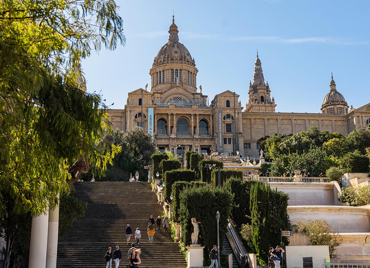 Grand view of the Museu Nacional d'Art de Catalunya (MNAC) in Barcelona, with its domed central tower and ornate facade rising above a wide staircase flanked by trimmed hedges and statues. Visitors are seen walking up the steps on a sunny day, surrounded by lush greenery.







