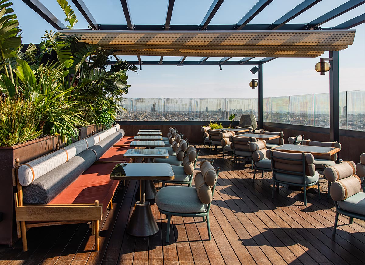 Stylish rooftop bar at Nobu Hotel in Barcelona, featuring sleek lounge seating with cushioned chairs and long upholstered benches under a modern pergola. The wooden deck is lined with lush green plants and glass railings that offer a panoramic view of the city skyline. Soft sunlight enhances the warm, inviting atmosphere of the open-air space.
